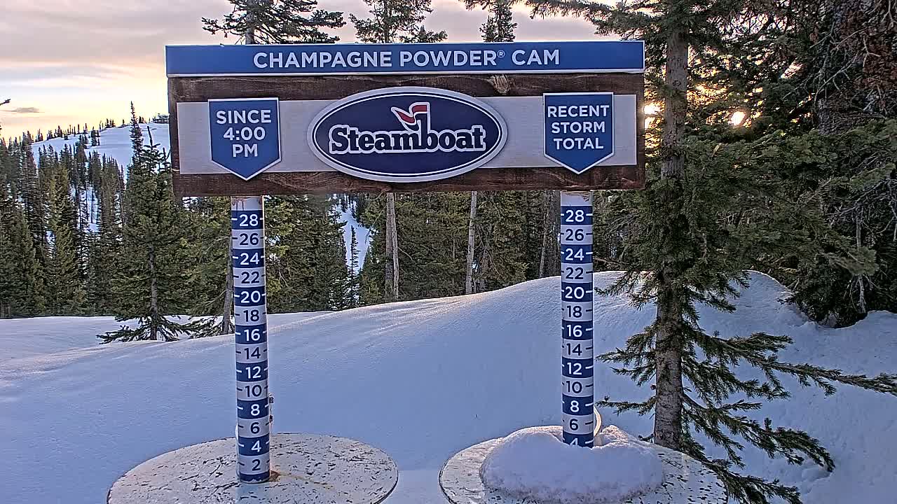 A snow depth gauge stands in a snowy landscape at sunset, surrounded by evergreen trees.