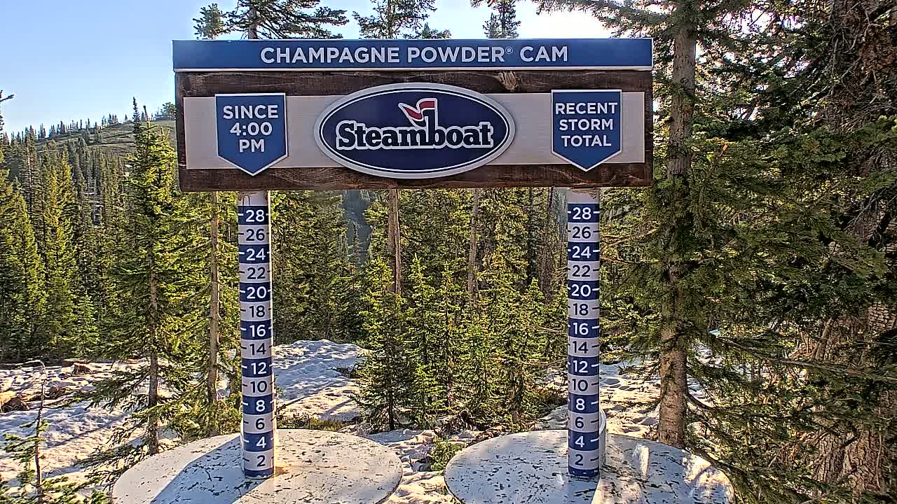 A snow depth measurement station shows recent snowfall in a sunny, mountainous area with evergreen trees.