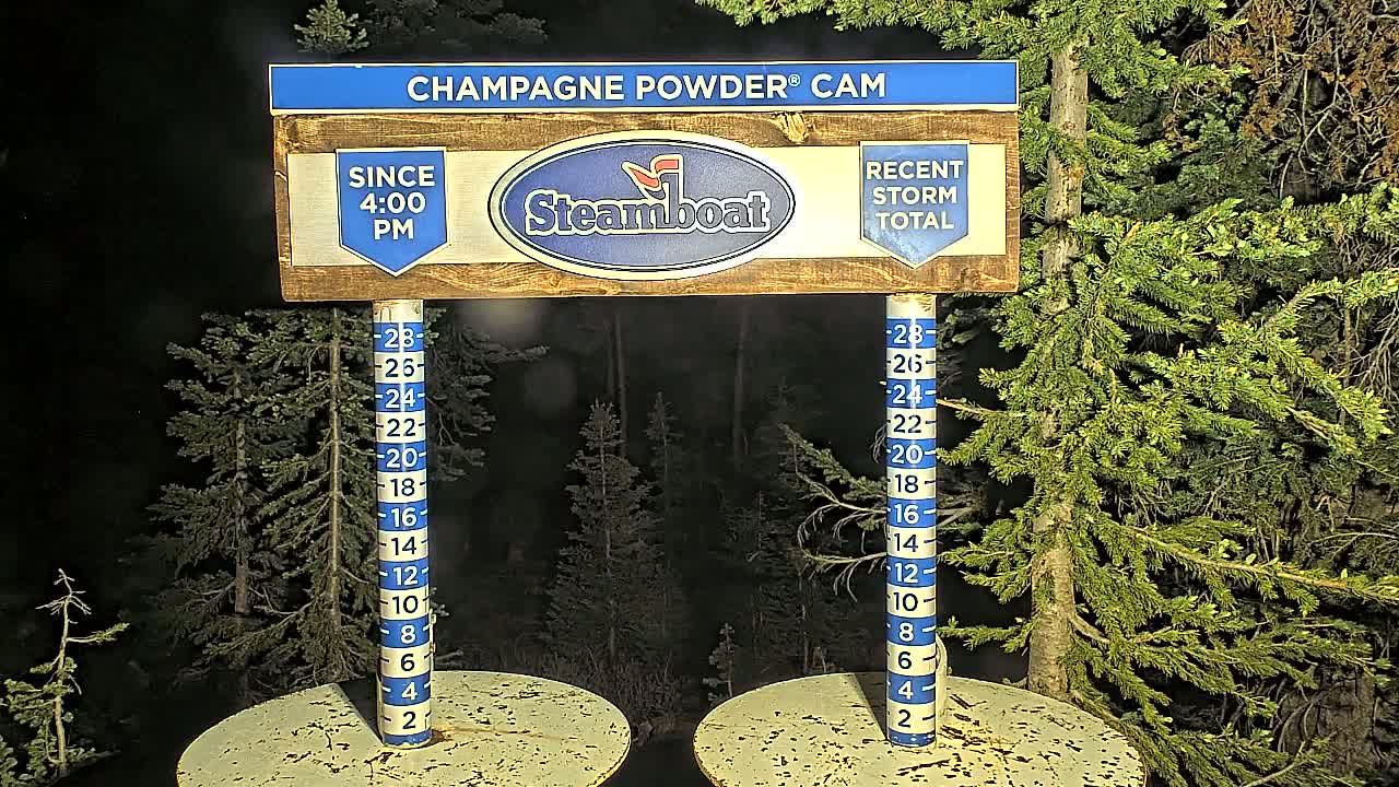 A nighttime snow depth gauge with a "Steamboat" logo sits amongst evergreen trees in snowy conditions.