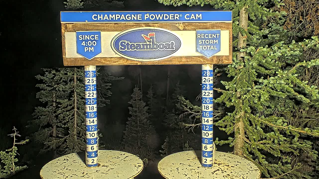 At night, a wooden sign displaying snowfall measurements stands between two round tables amidst evergreen trees.