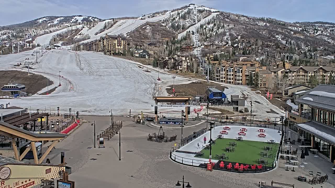 A mostly snow-covered ski slope sits below a mountain village under a partly cloudy sky.