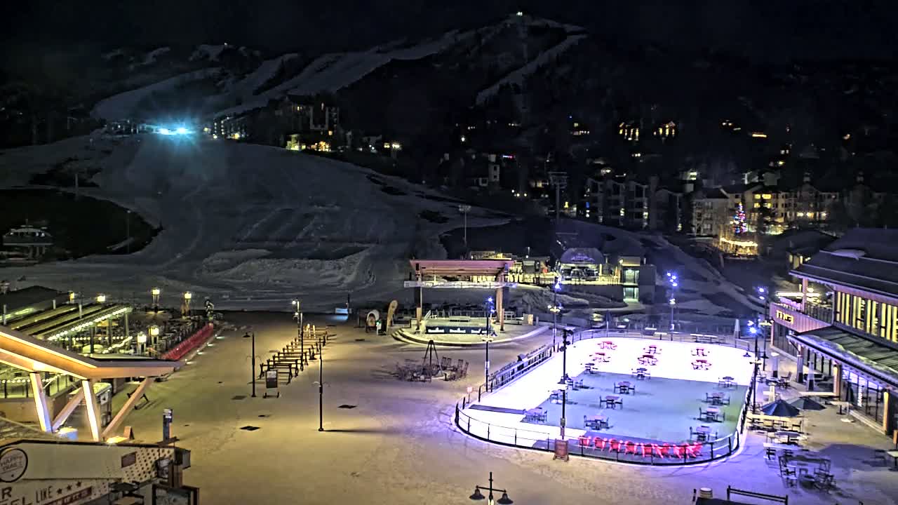 An outdoor ice rink illuminated at night is surrounded by snow-covered ground, buildings, and a ski slope in the background.