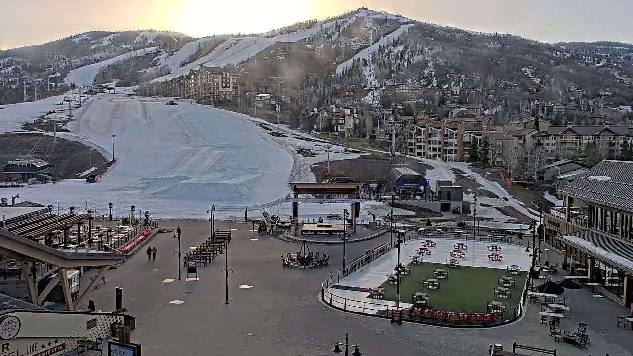 A snow-covered ski resort at sunset, with a plaza featuring an ice rink and outdoor seating below, and residential buildings and ski slopes on the hillside above.