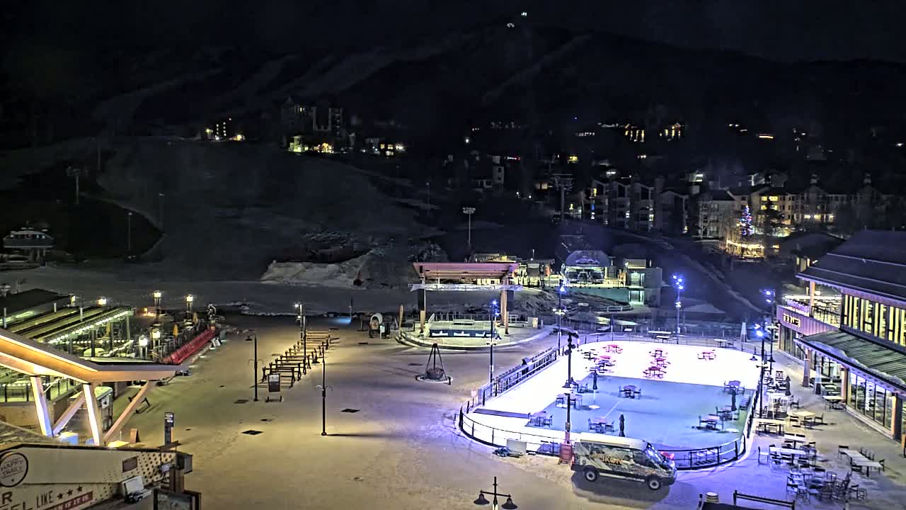 An illuminated ice rink sits in a snowy plaza at night, surrounded by buildings and ski slopes on a hillside under a clear, dark sky.
