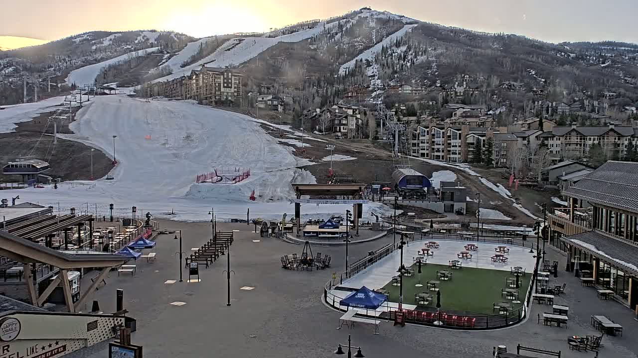 A snow-covered ski slope and base area with buildings and outdoor seating areas are visible at sunset.