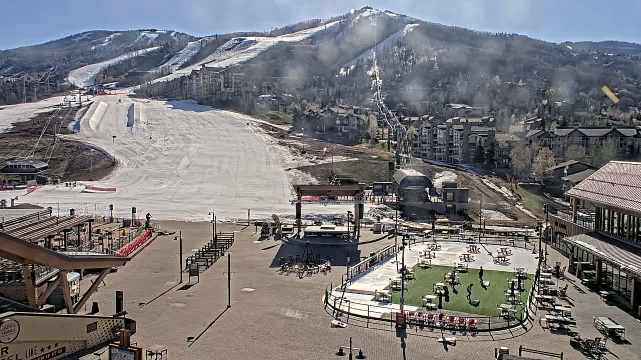 A sunny day reveals a snow-covered ski slope overlooking a plaza with an ice rink and outdoor seating, nestled among buildings in a mountain resort town.