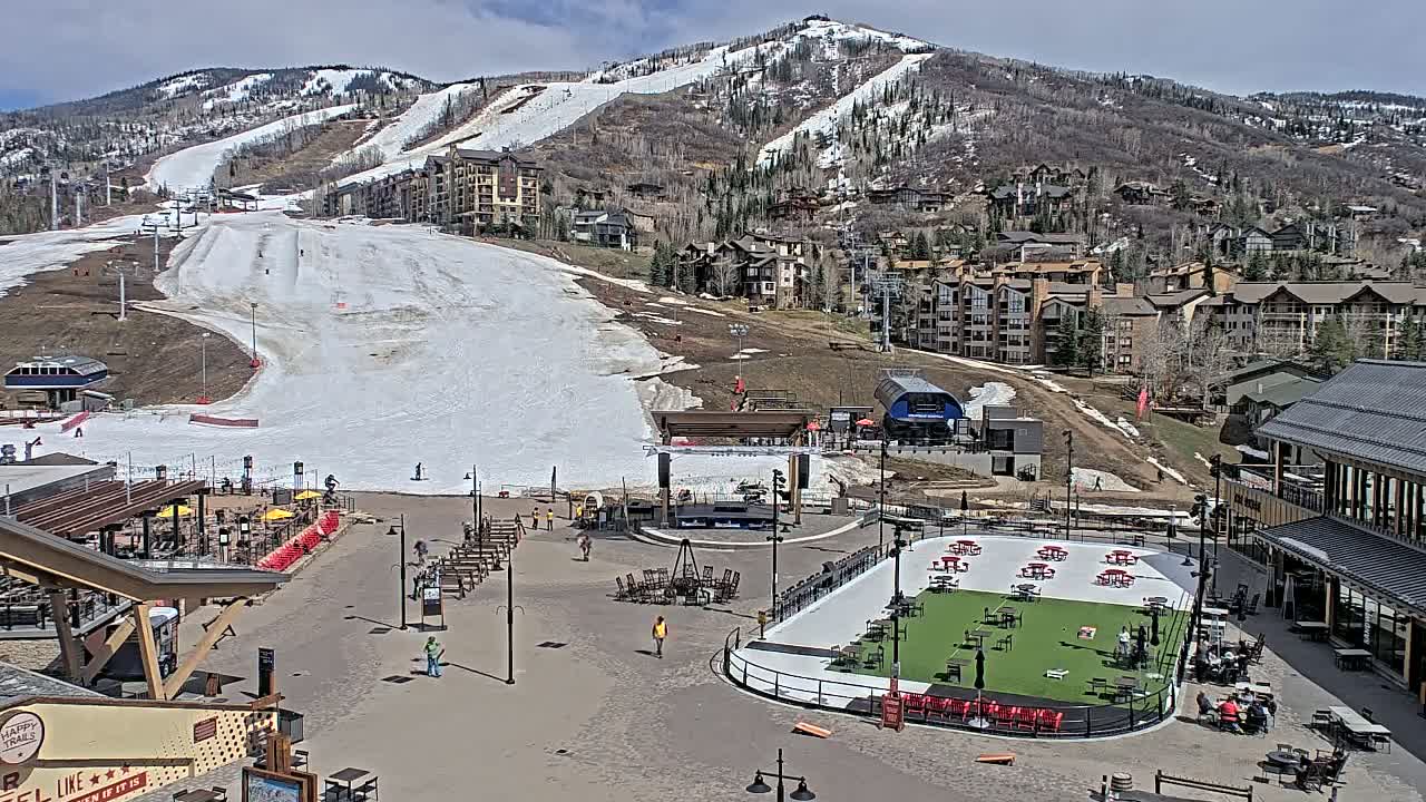 A mostly sunny day at a ski resort base area shows a snow-covered ski slope on a mountain behind a plaza with several outdoor seating areas and a small, artificial turf field.