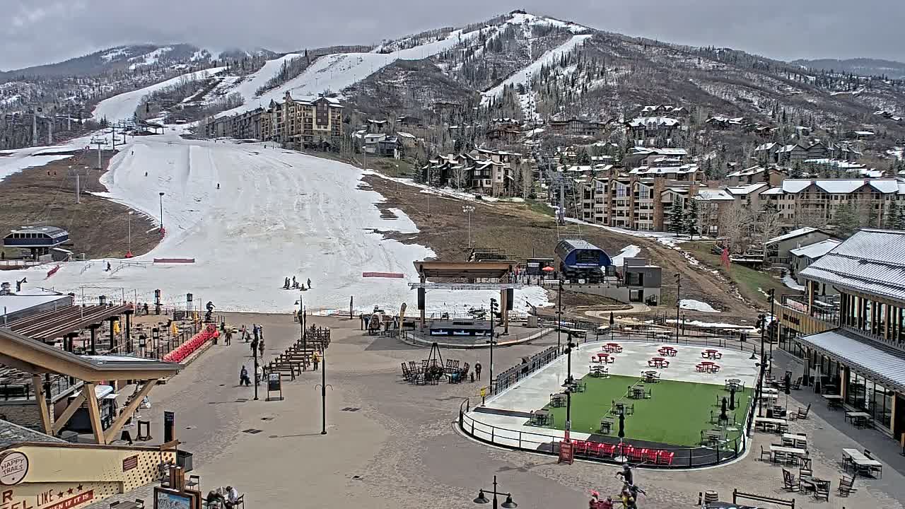 A snow-dusted ski slope overlooks a village base area with outdoor seating and a partially melted ice rink, under overcast skies.