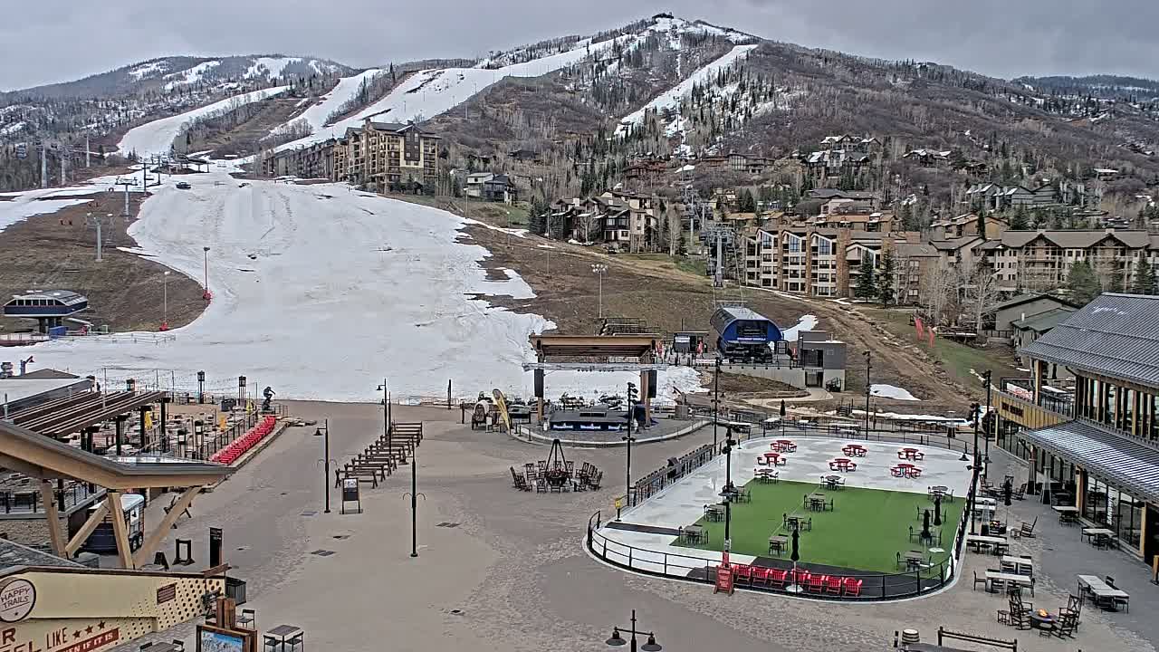 An outdoor plaza with an ice rink and a stage sits below a ski resort with snow-covered slopes and buildings on a cloudy day.