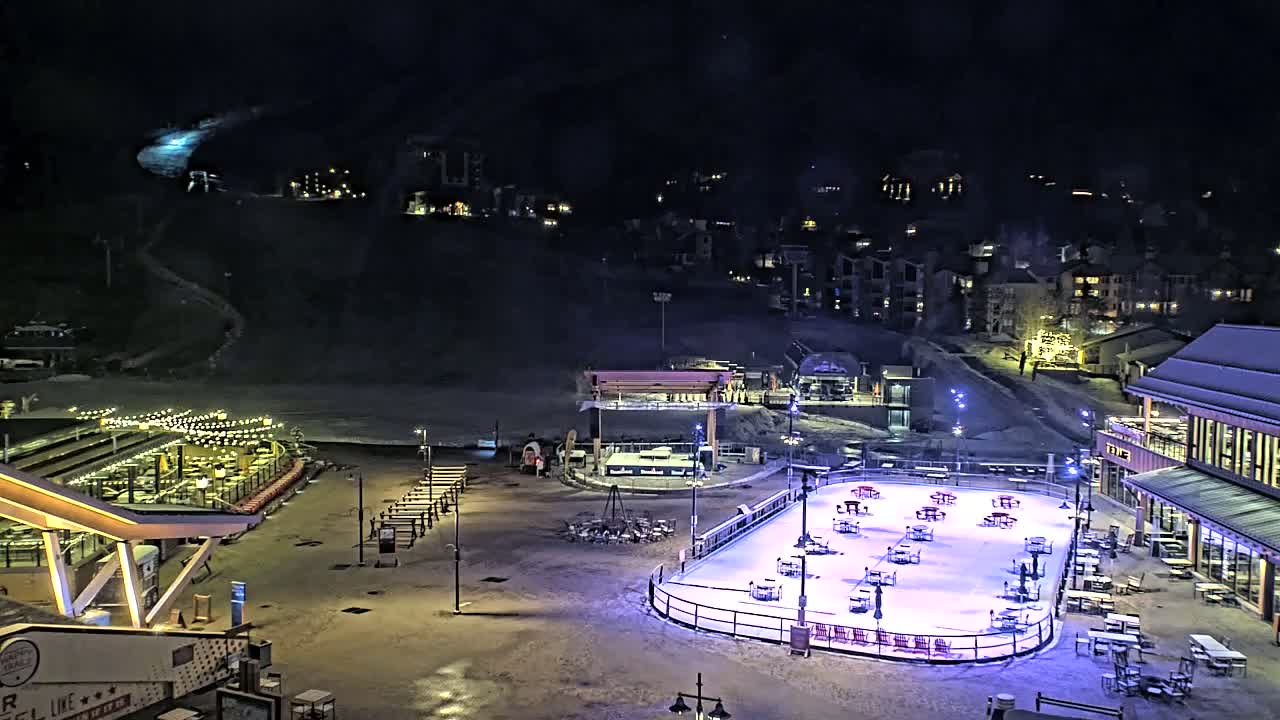An outdoor ice rink is illuminated at night, surrounded by buildings, a restaurant, and a snow-covered hillside under a dark sky.