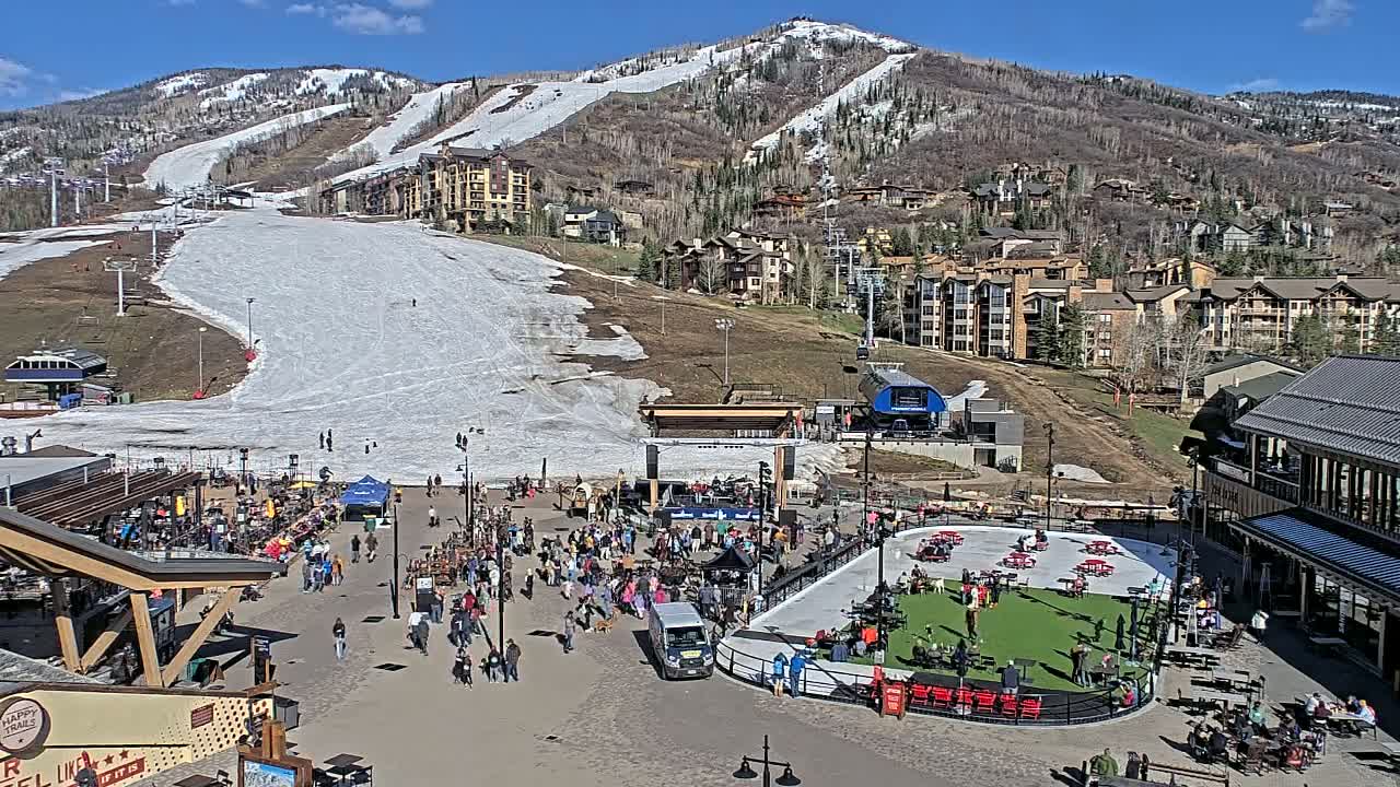 A sunny day at a ski resort base area shows a large crowd gathered around an outdoor stage and ice rink, with snow-covered slopes and buildings in the background.