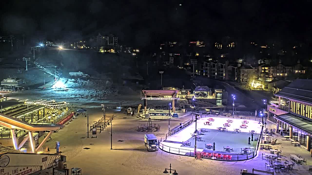 A nighttime aerial view of a snow-covered ski resort base area, featuring a lit-up ice skating rink, outdoor seating areas, and buildings, under a dark sky.