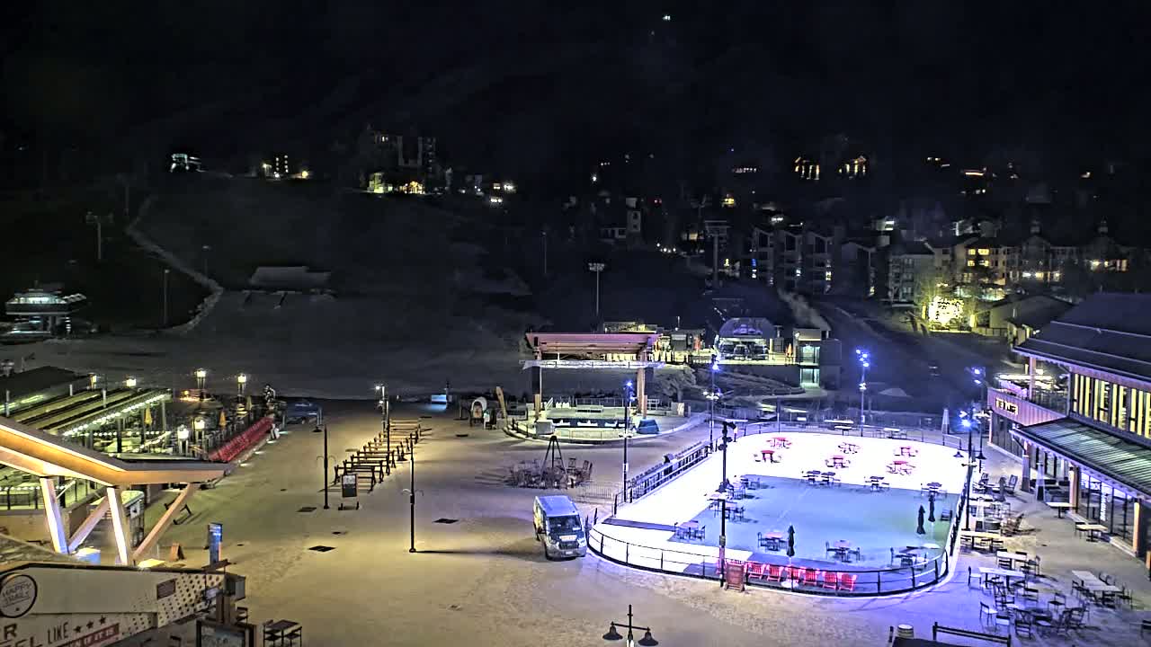 A nighttime view of a snow-covered plaza with an ice rink, buildings, and a ski slope in the background.