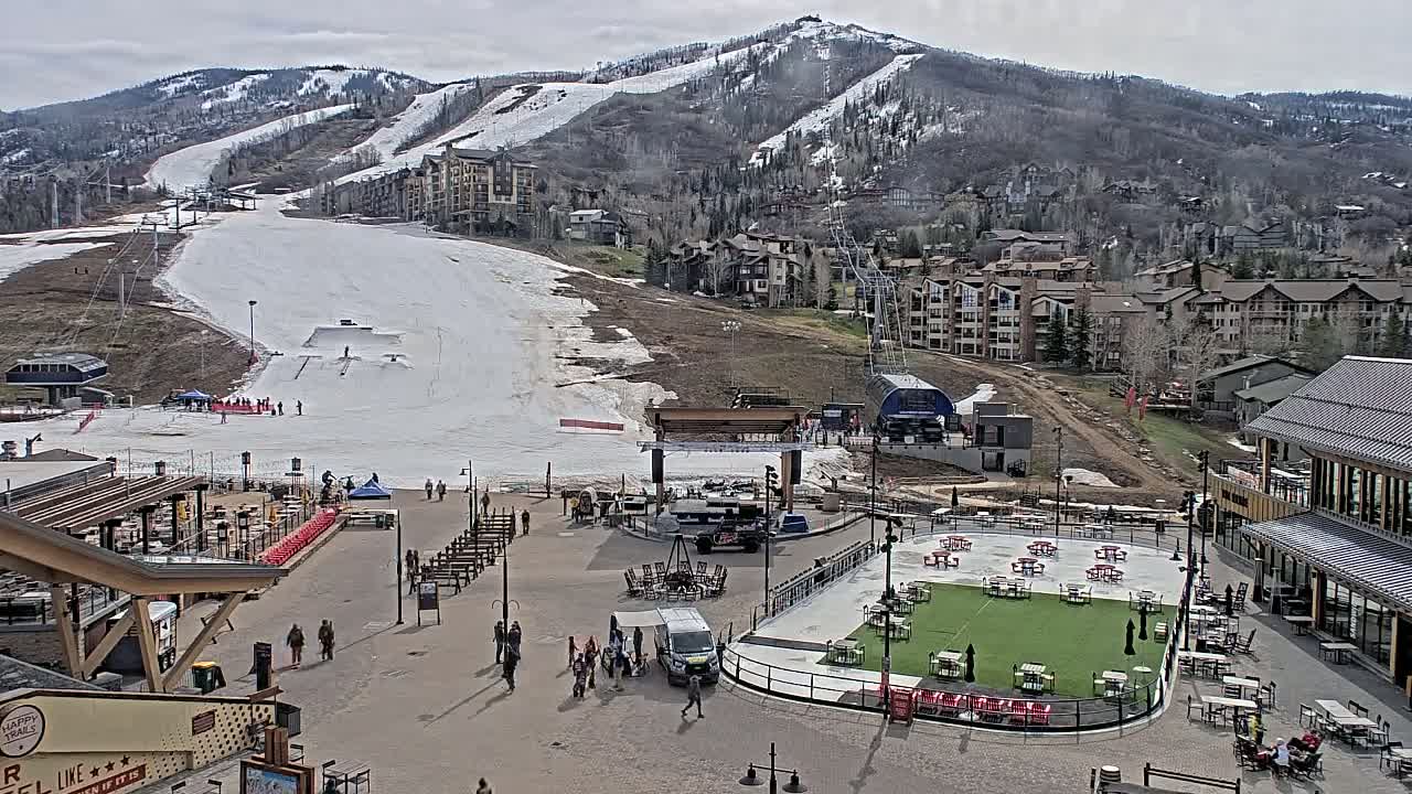 A partially snow-covered ski resort base area with several buildings, outdoor seating areas, and people, under an overcast sky.