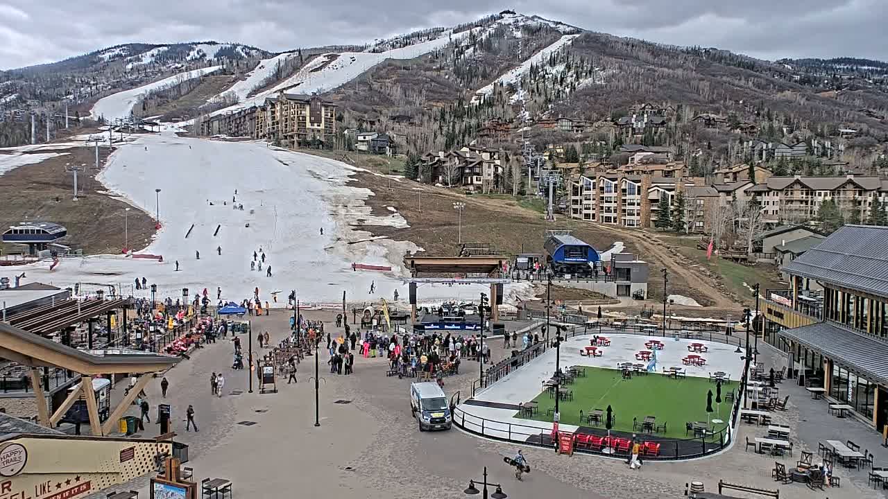 A partially snow-covered ski slope overlooks a village square with a crowd of people gathered near an outdoor stage and an ice rink under an overcast sky.