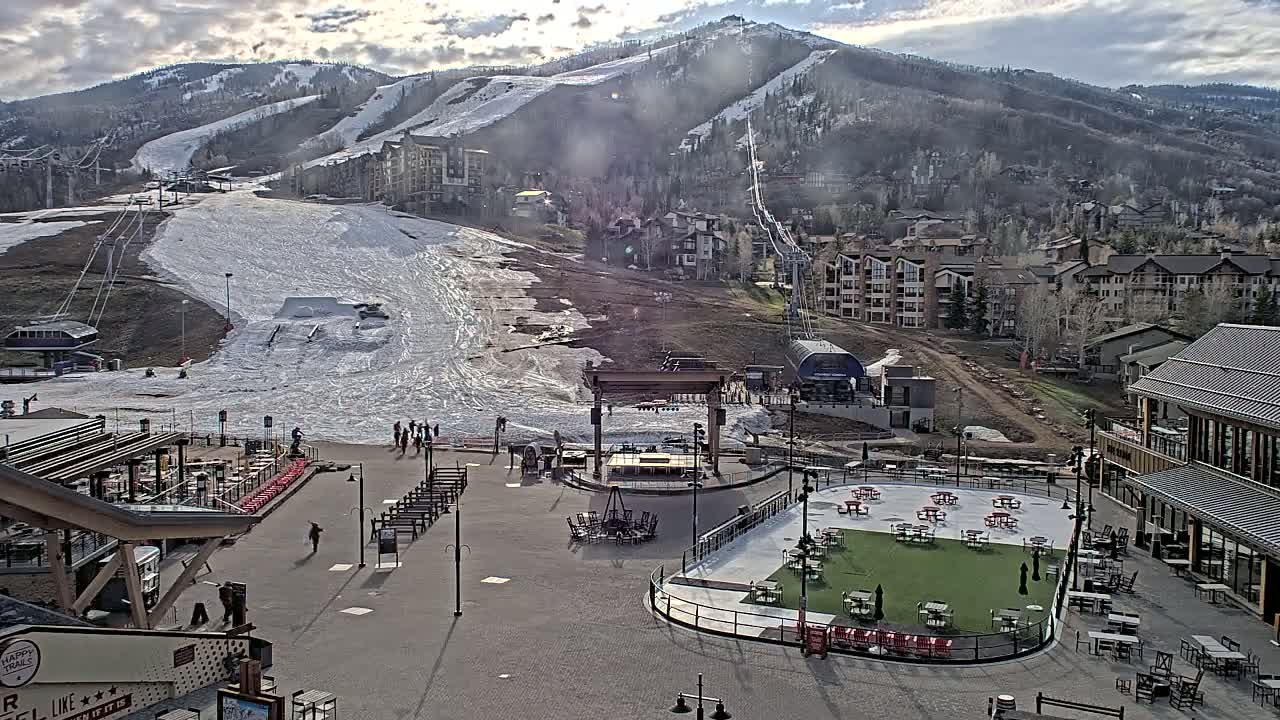 A mostly sunny day shows a partially snow-covered ski slope with buildings and an outdoor plaza below, which includes picnic tables and a stage.