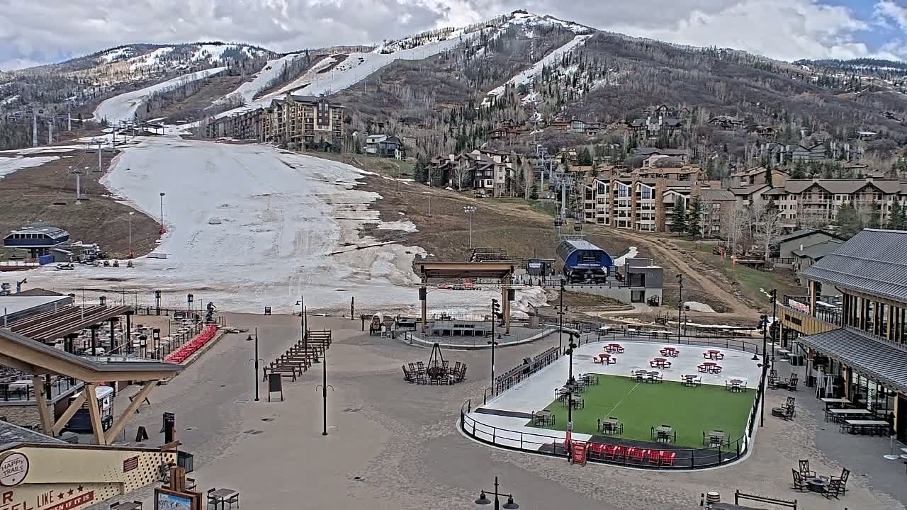An outdoor plaza with a stage, fire pit, and artificial turf field is situated below a snow-covered ski slope and mountainside village under a partly cloudy sky.