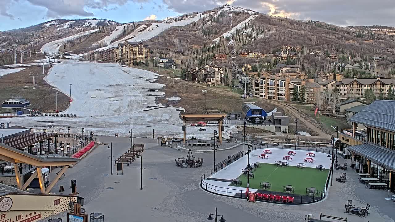 A mostly snow-covered ski slope and base area with buildings and an outdoor ice rink, under a partly cloudy sky.