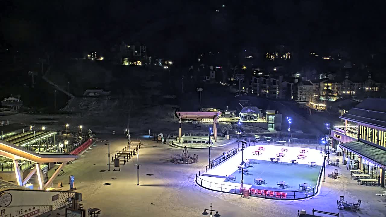 A nighttime view of a snow-covered outdoor ice rink surrounded by buildings and seating areas under a clear, dark sky.