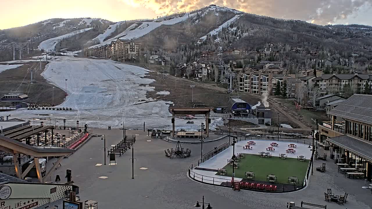 A snow-covered ski slope descends towards a village of buildings and a plaza with an outdoor rink and seating areas at sunset.