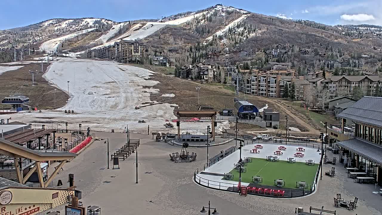 A mostly snow-free ski slope and resort village are visible under a clear blue sky, with a paved plaza area containing an outdoor stage and seating.