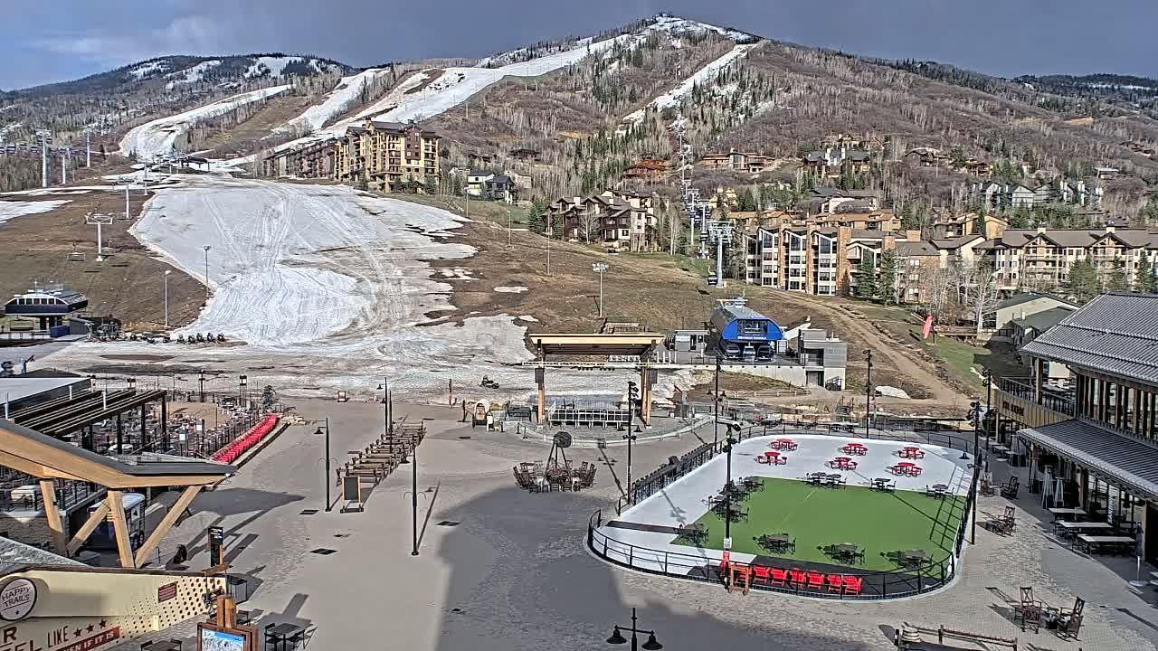 A mostly sunny day reveals a ski resort base area with a partially snow-covered ski slope, buildings, and an outdoor ice rink.