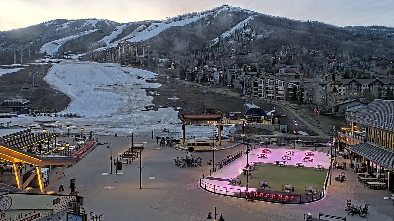 A snow-covered ski slope and village are visible under a cloudy sky, with an ice rink and outdoor seating areas in the foreground.