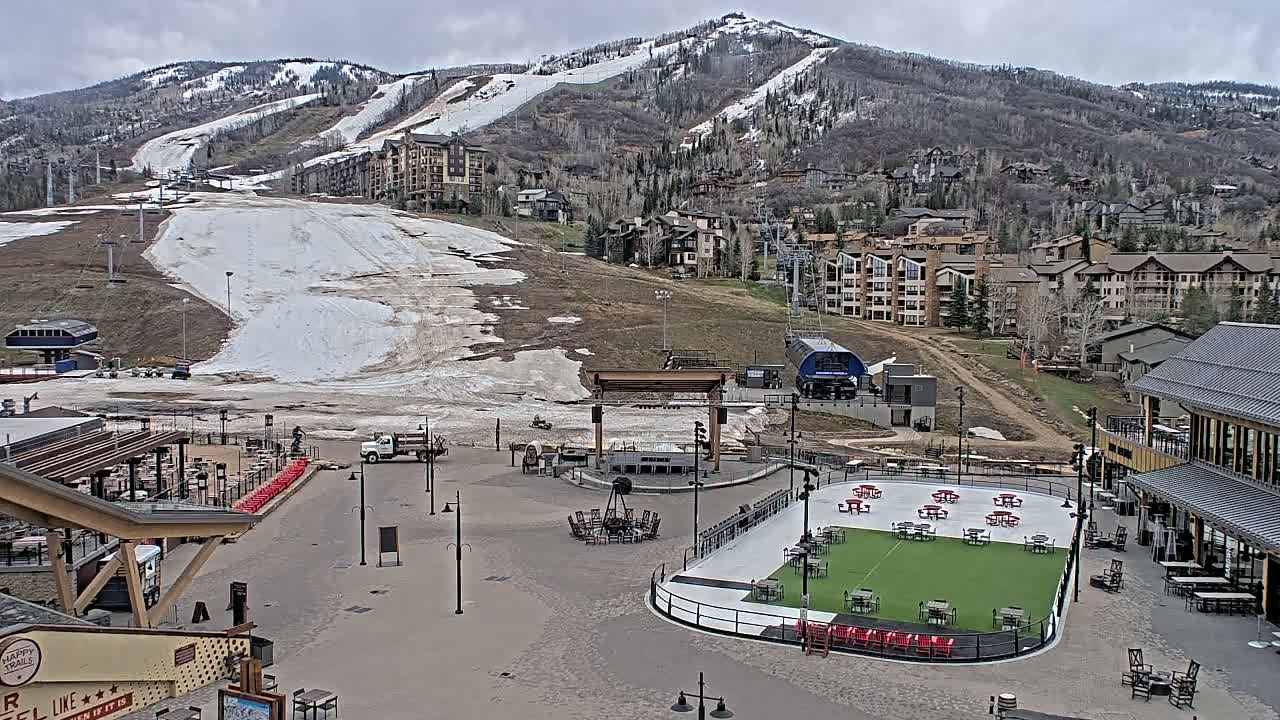 A mostly clear day shows a snow-covered mountain slope above a village with buildings and outdoor seating areas around a paved plaza; some melting snow is visible on the lower slope.