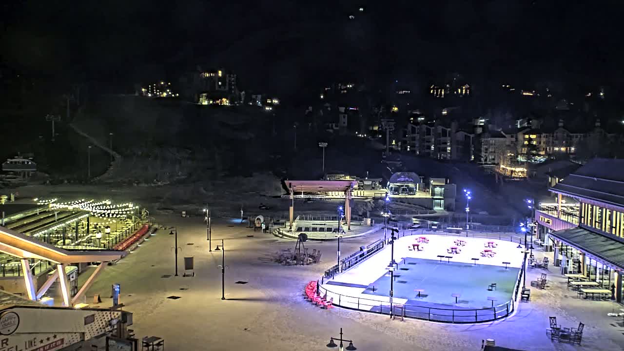 A snow-covered plaza at night, illuminated by artificial light, features a lit ice rink surrounded by picnic tables and buildings, with a hill and buildings in the background.
