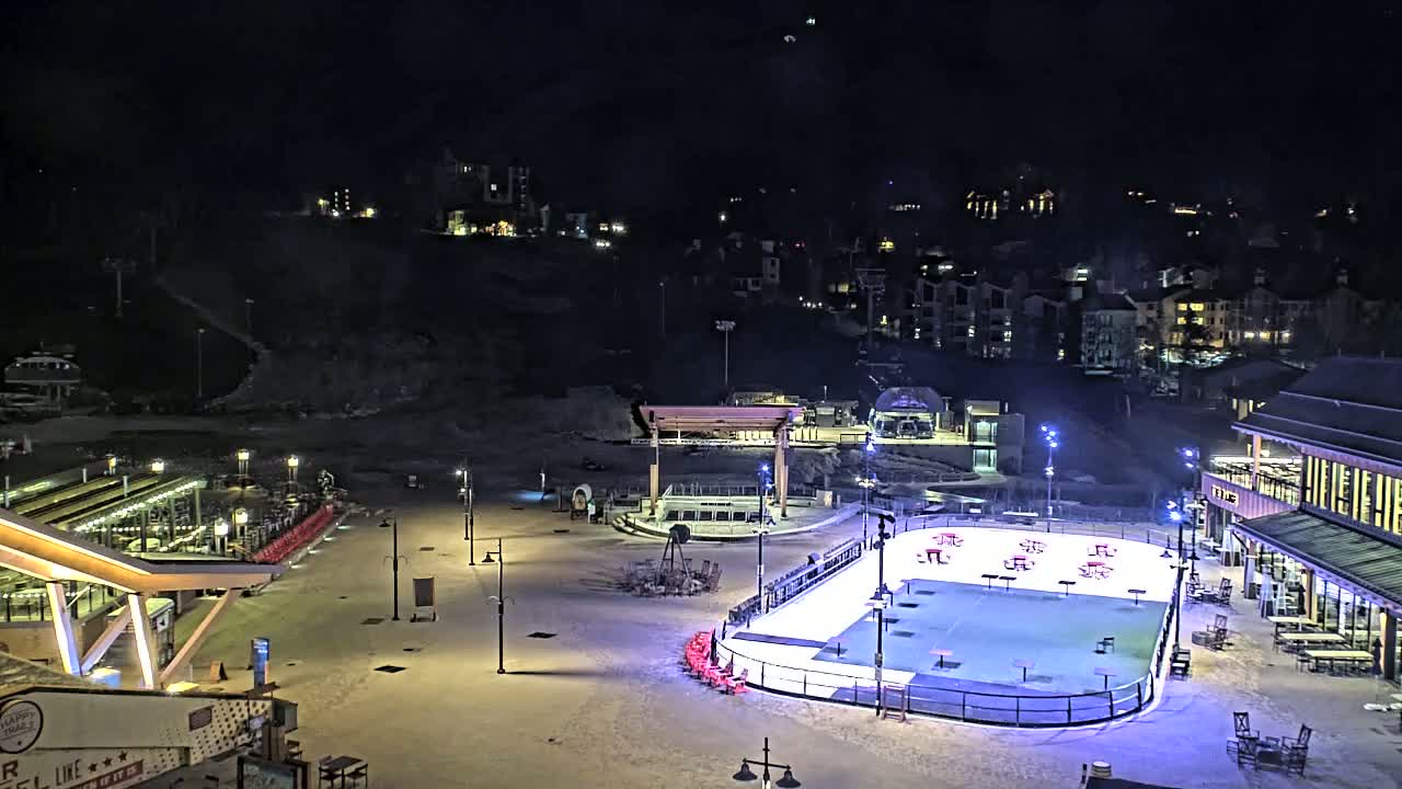 An outdoor ice rink, illuminated at night, is situated in a snow-covered plaza near buildings and a ski lift, under a dark, clear sky.
