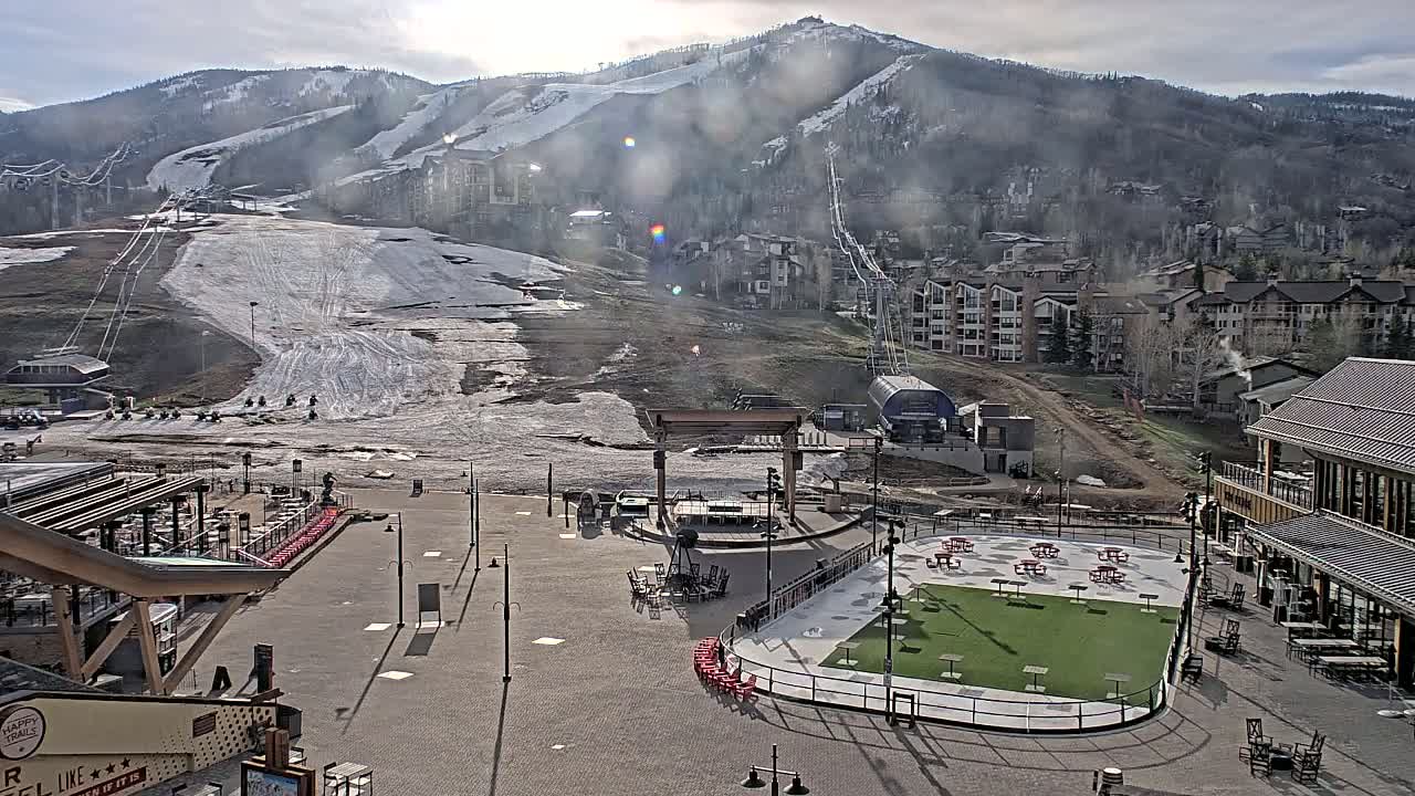 A partially snow-covered ski slope and base village are visible under a mostly cloudy sky, with a paved plaza containing picnic tables and a small artificial turf area in the foreground.