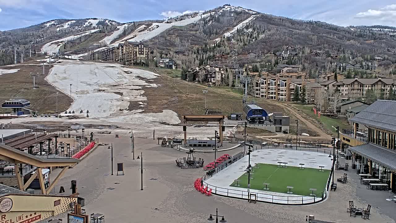 A mostly snow-free ski resort base area with a partially melted ice rink, surrounded by buildings and a mountain with patches of snow under a partly cloudy sky.