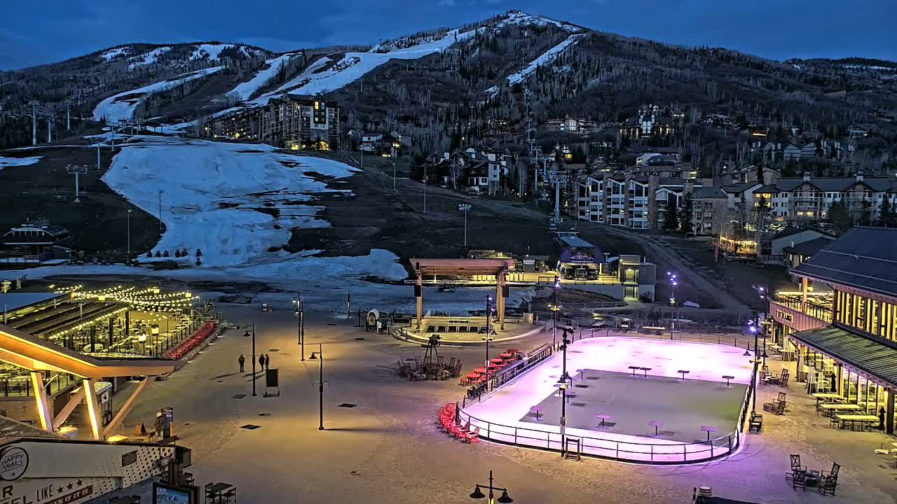 An outdoor ice rink is illuminated at night, situated at the base of a snow-covered mountain with buildings and ski lifts visible in the background.