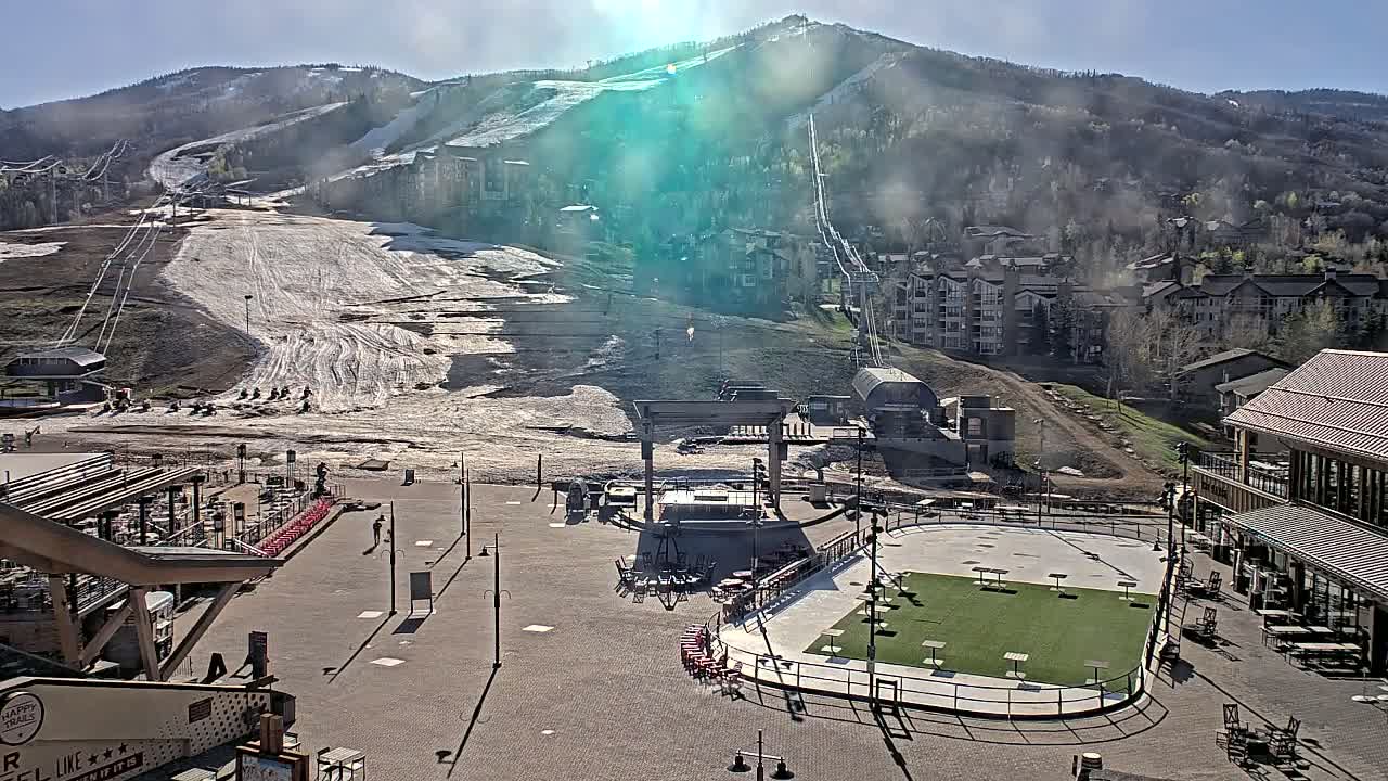 A sunny day reveals a snow-covered ski slope, a village of buildings nestled in the mountains, and a plaza below with an outdoor stage and artificial turf area.