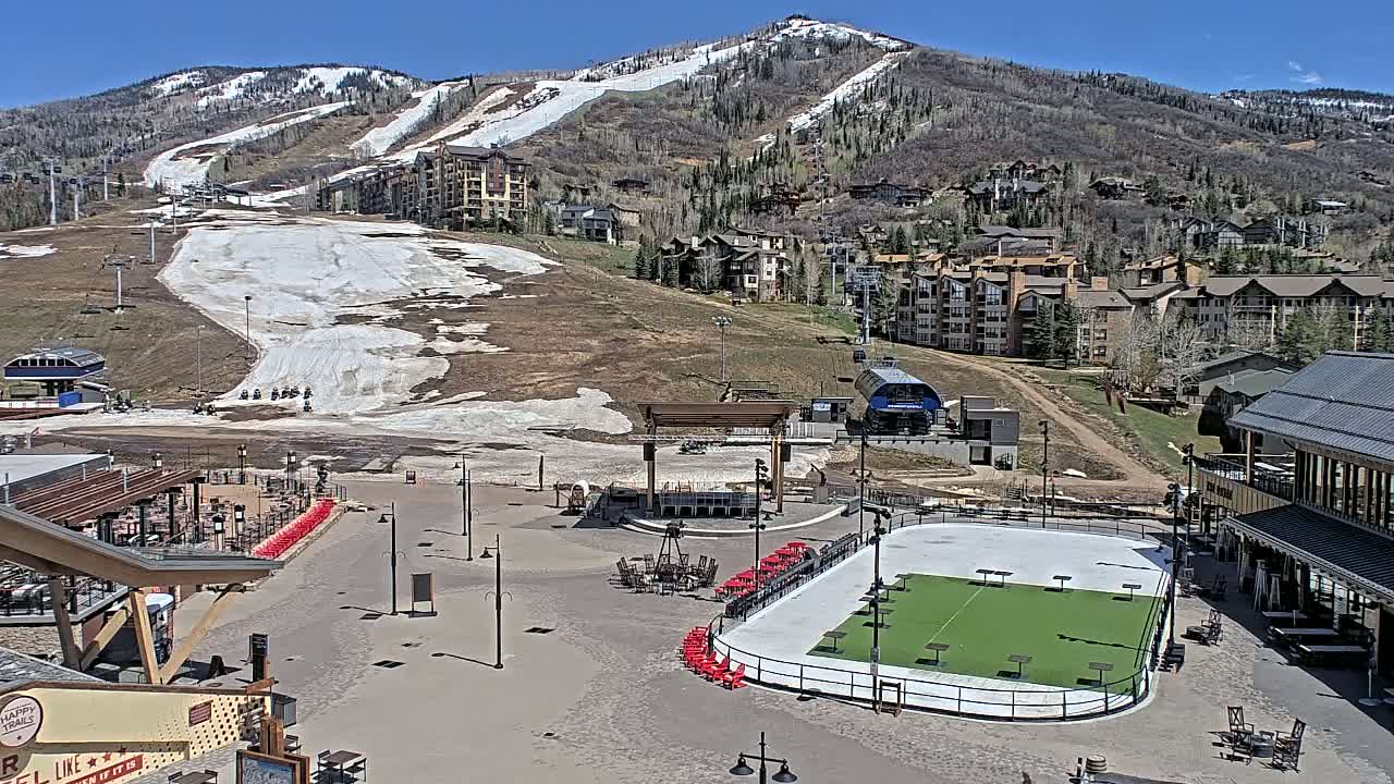 A sunny day reveals a mountainside ski resort with melting snow patches on the slopes, a village of buildings below, and a plaza containing an outdoor ice rink that has been converted into a turf area.