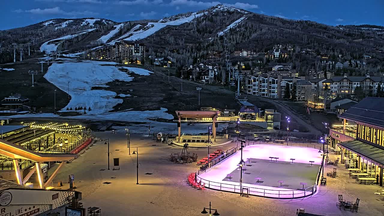 An illuminated outdoor ice rink sits in a snow-covered plaza at the base of a snow-dusted mountain at dusk, with buildings and ski lifts visible on the mountainside.