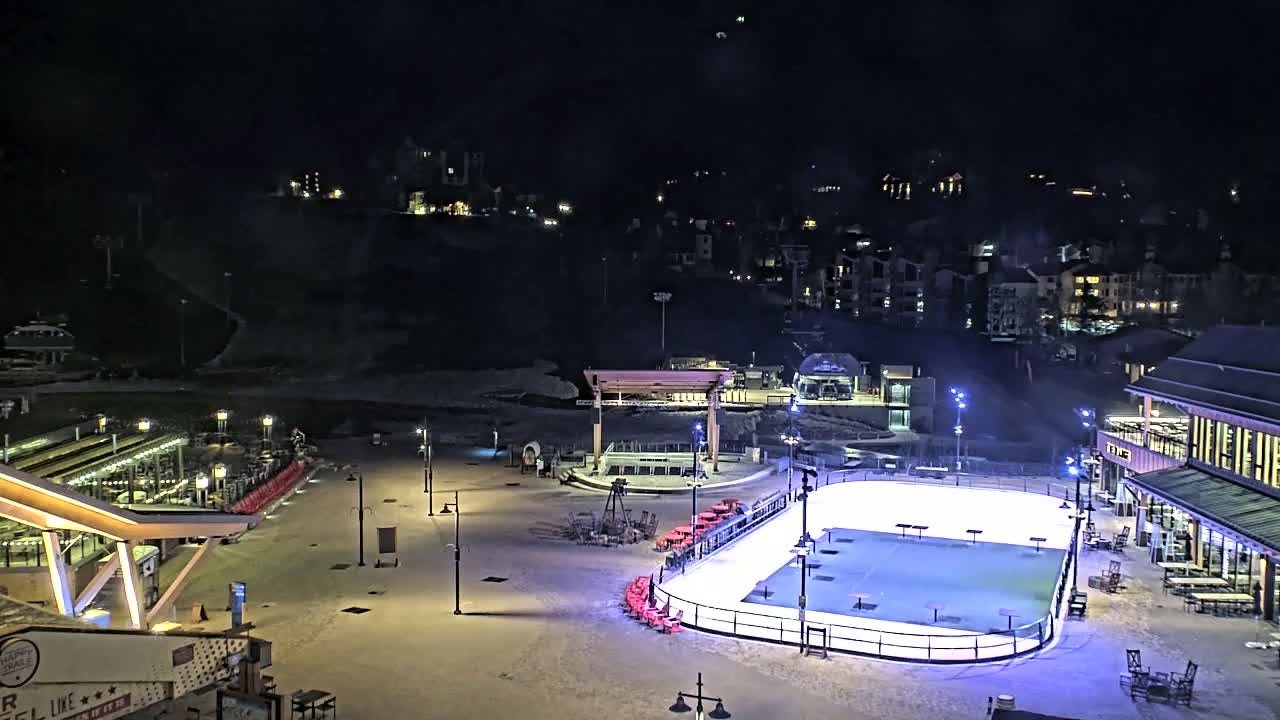 An outdoor ice rink is illuminated at night, surrounded by buildings and snow-covered ground under a clear, dark sky.