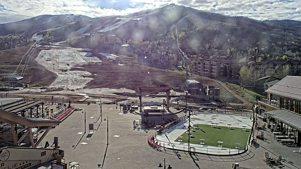 A mostly sunny day reveals a partially snow-covered ski slope and village base area, featuring an outdoor plaza with a stage and artificial turf playing field.