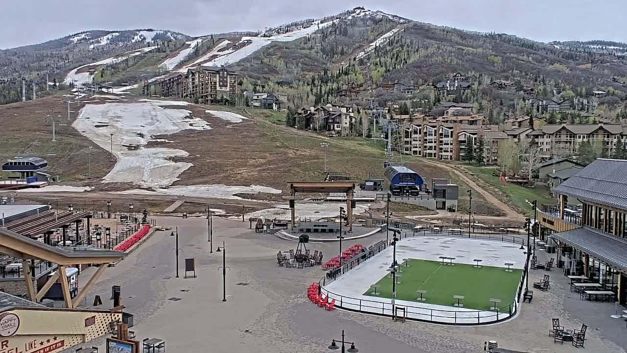 A mostly clear day reveals a ski resort base area with a paved plaza, an empty ice rink, and buildings, all situated below a mountain with patches of snow and spring foliage.