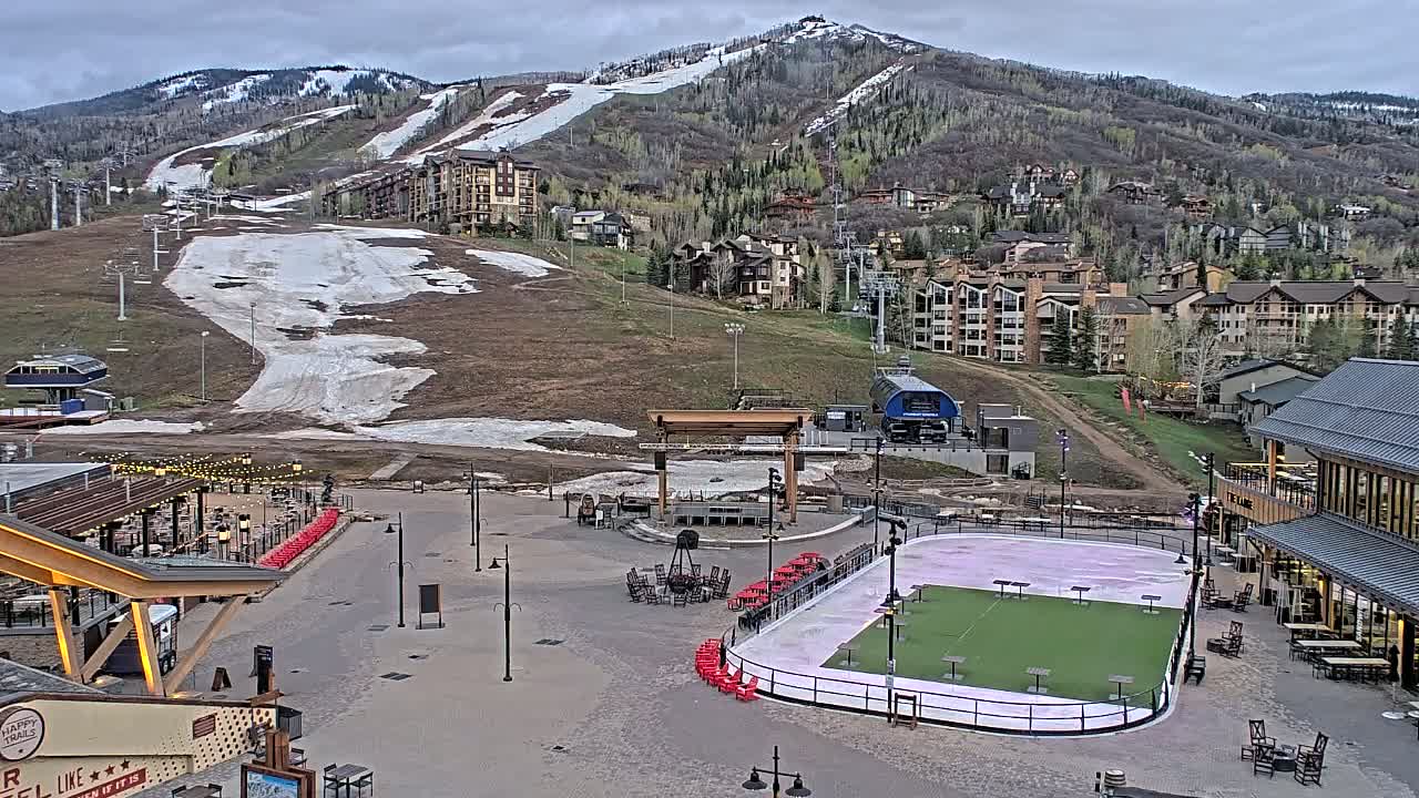 A mostly clear day reveals a mountain resort with a partially snow-covered ski slope, buildings, and a paved plaza containing an ice rink and outdoor seating.