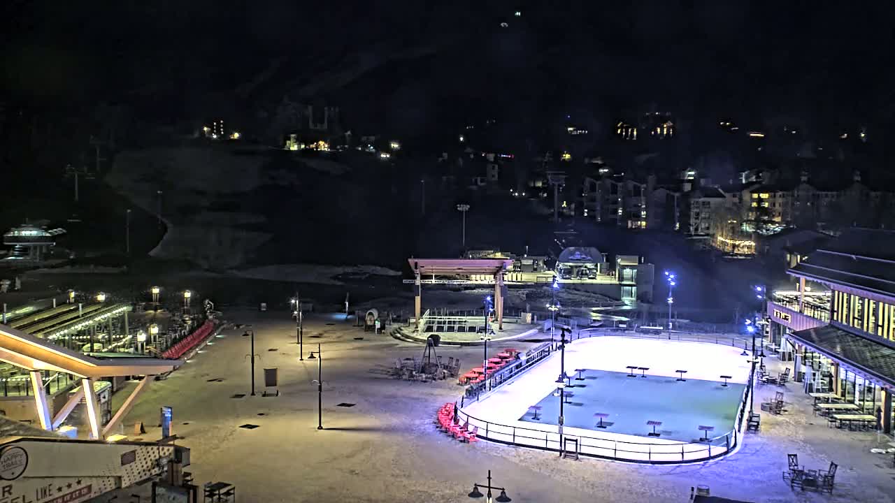 An outdoor ice rink is illuminated at night, surrounded by snow-covered ground, buildings, and ski lift infrastructure.