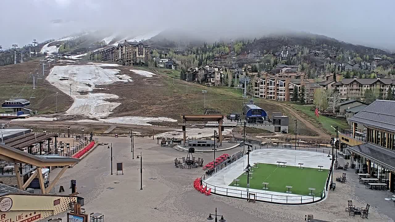 A mostly snow-free ski resort base area with a partially melted ice rink, surrounded by buildings and a mountain partially covered in fog.
