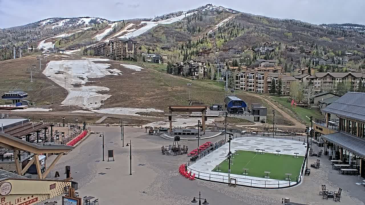 A mostly clear day reveals a mountainside ski resort with a partially snow-covered slope, buildings, a plaza with an outdoor ice rink, and seating areas.