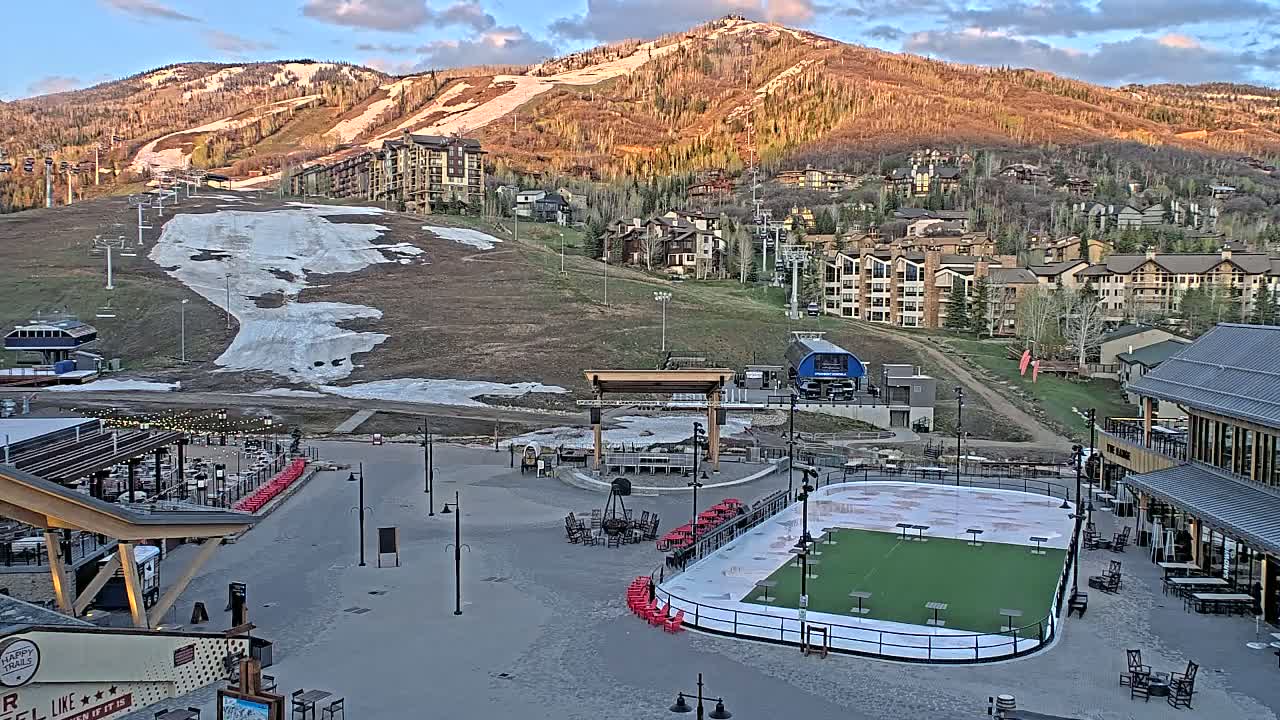 An outdoor plaza with a skating rink and several buildings is nestled at the base of a mountain with ski slopes showing patches of snow under a partly cloudy sky.