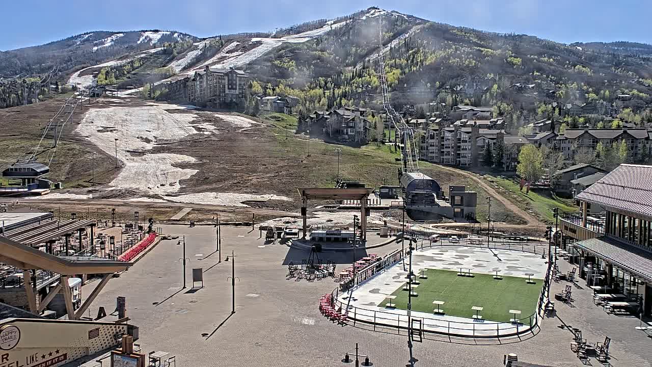 A sunny day reveals a mountainside resort village with a partially snow-covered ski slope, buildings, and a plaza with an empty ice rink.