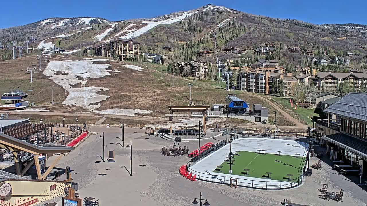 A sunny day reveals a mountainside resort with a partially snow-covered ski slope, residential buildings, an outdoor plaza containing a stage and an ice rink, and a restaurant.