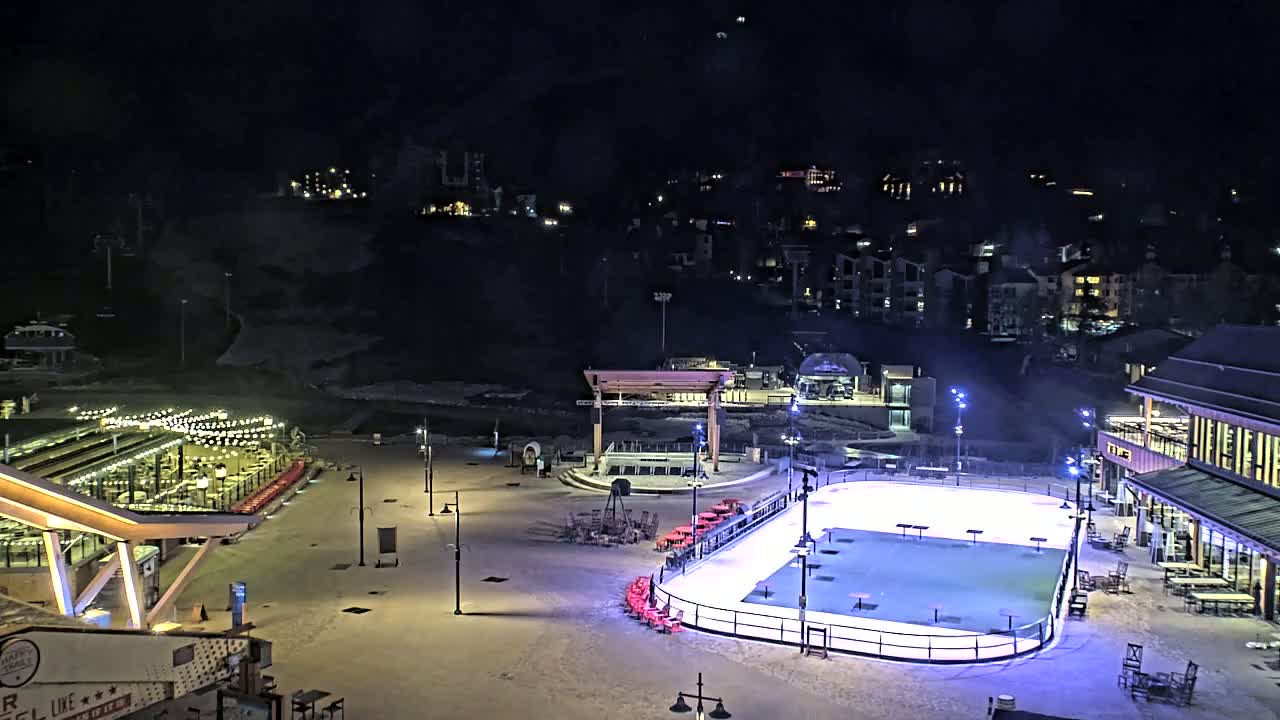 An outdoor ice rink is illuminated at night, surrounded by buildings and snow-covered ground, under a dark sky.