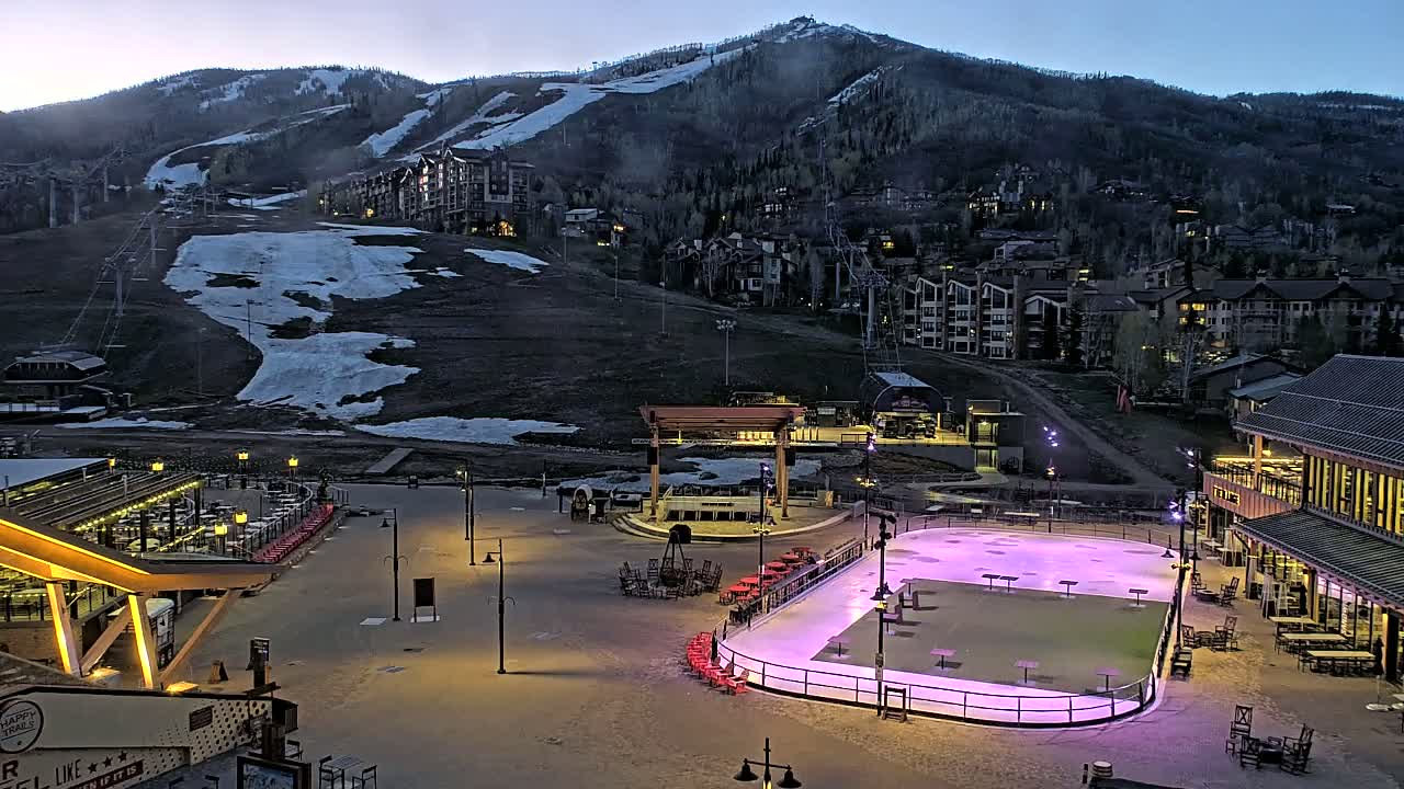 An outdoor ice rink illuminated at dusk, nestled in a mountain resort town with snow-dusted slopes visible in the background.