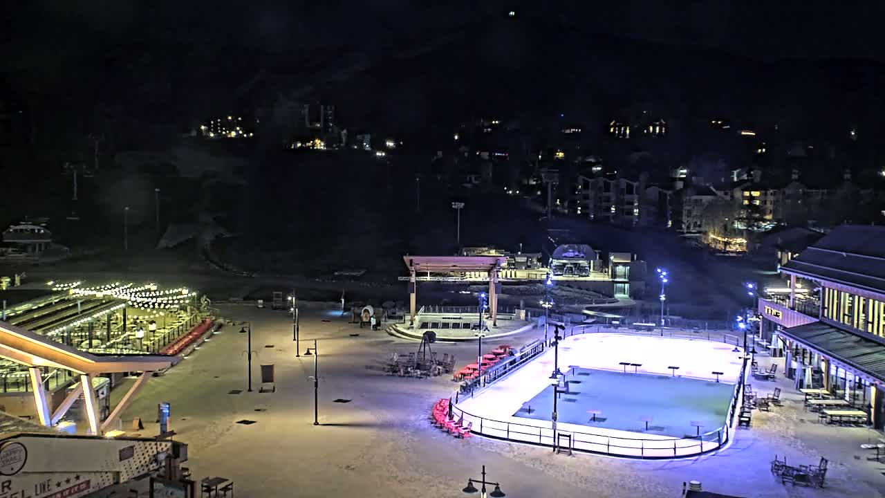 A snow-covered plaza at night, illuminated by artificial lights, features a lit ice rink, outdoor seating areas, and buildings in the background.
