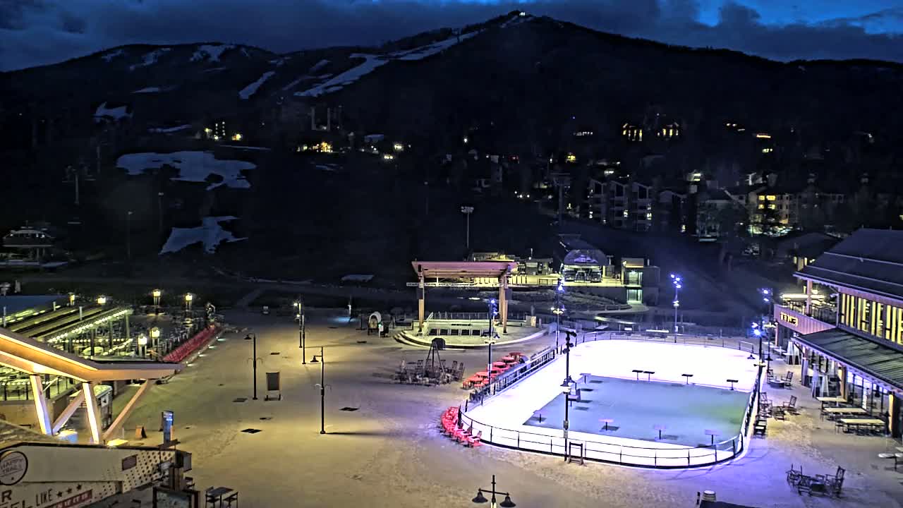 An outdoor ice rink is illuminated at night, situated below a snow-covered mountain with buildings and lights scattered across the landscape under a dark, cloudy sky.